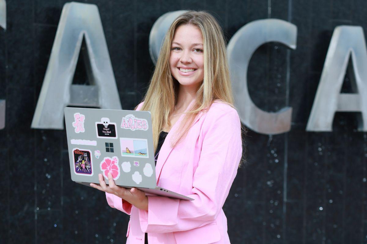 Smiling woman in a pink blazer holds a sticker-covered laptop outside.